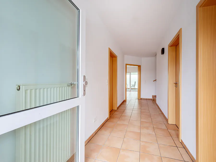 Hallway view with terracotta tile floor, white walls, and light wood doors. A white framed glass door is ajar on the left.