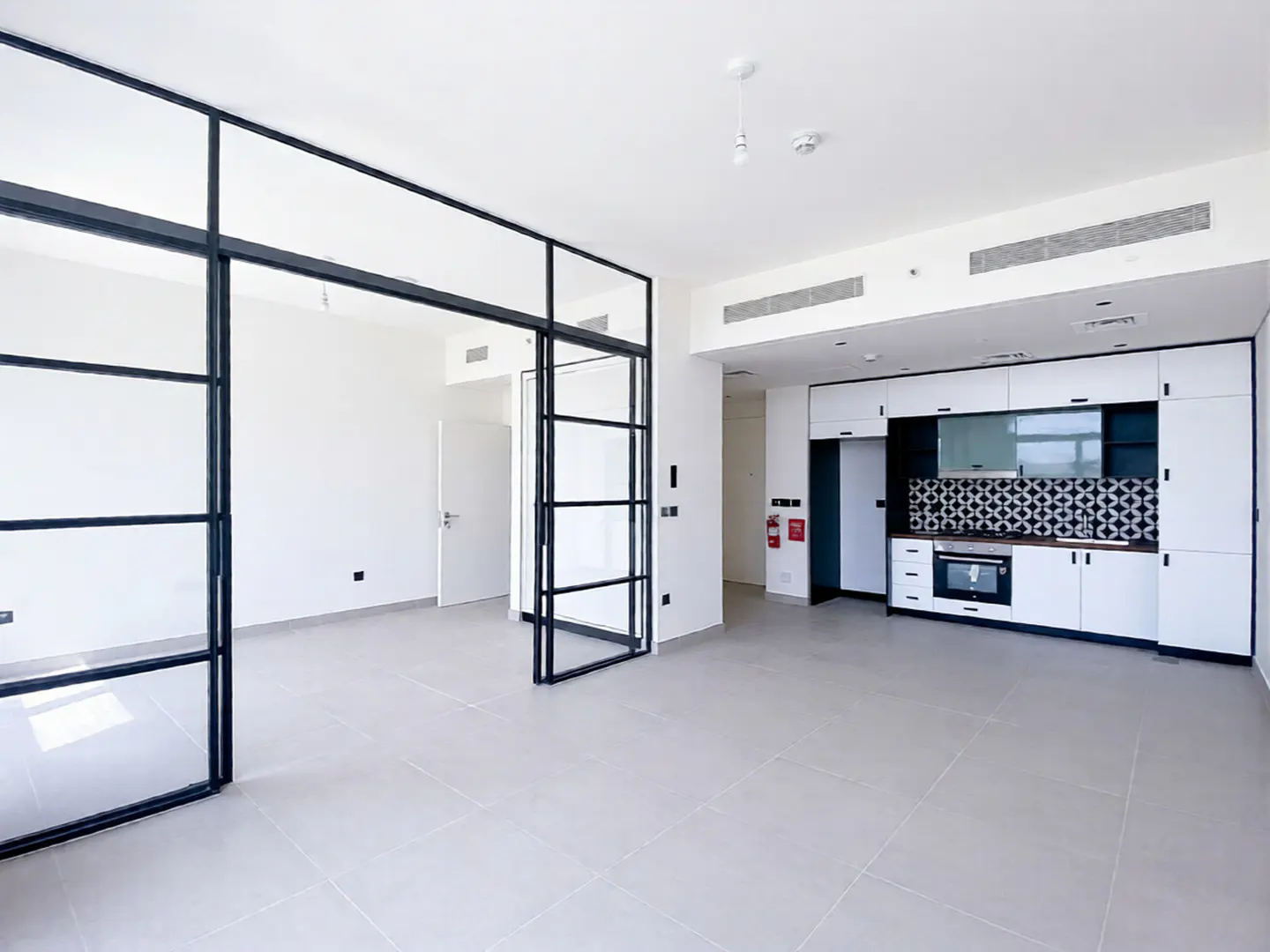 Bright, modern apartment interior with white walls, gray tile floor, and black-framed glass doors. A white kitchen with black accents is visible.