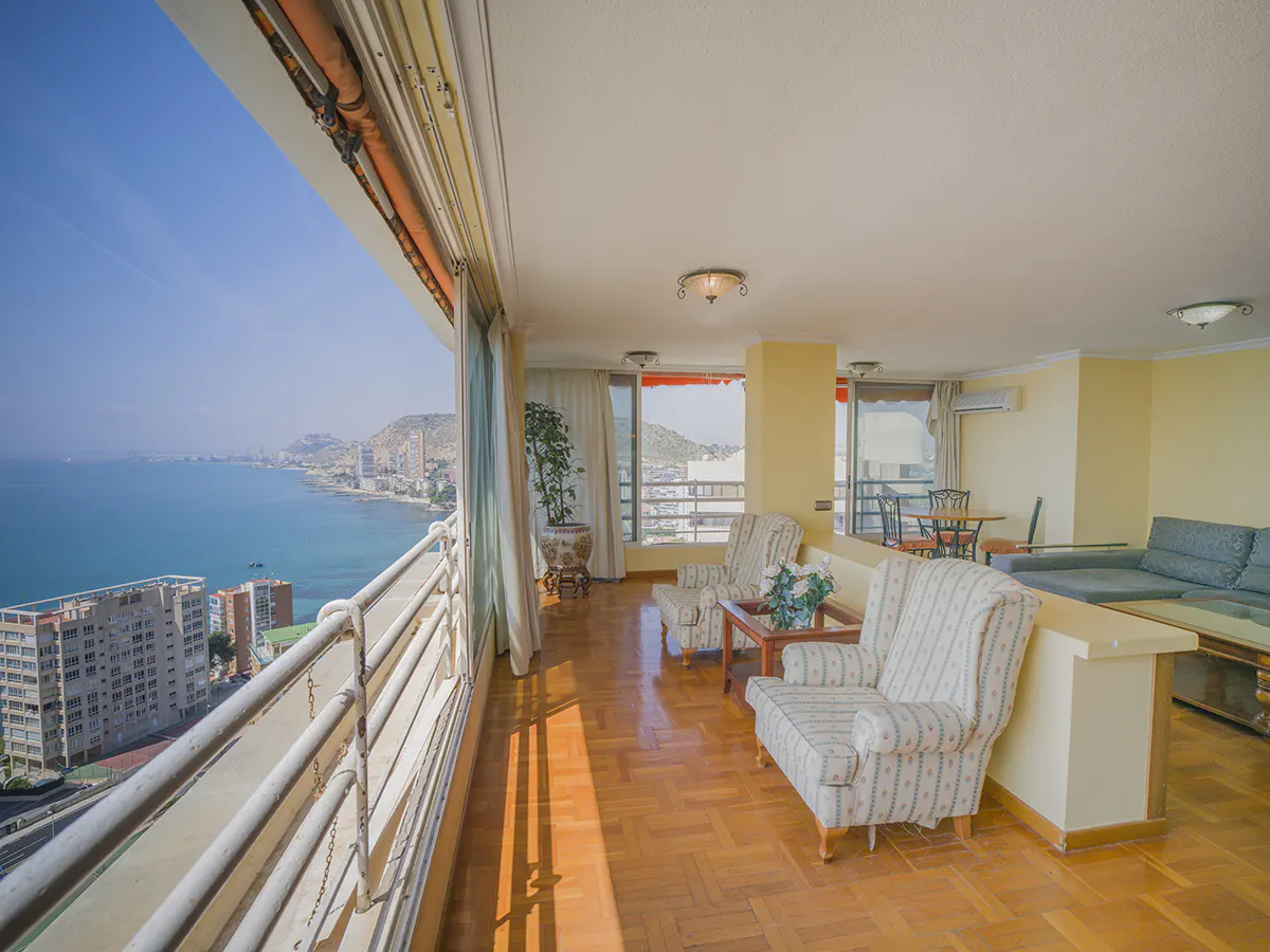 Bright apartment interior with a balcony overlooking the ocean. Armchairs, a sofa, and wood floors are visible.