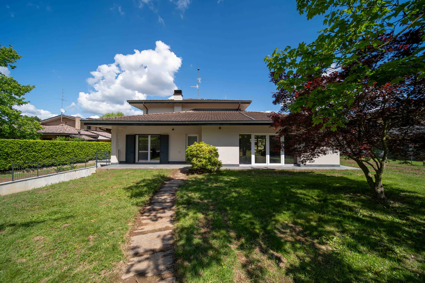 Exterior view of a one-story house with a brown roof, white walls, and a green lawn under a blue sky with white clouds.