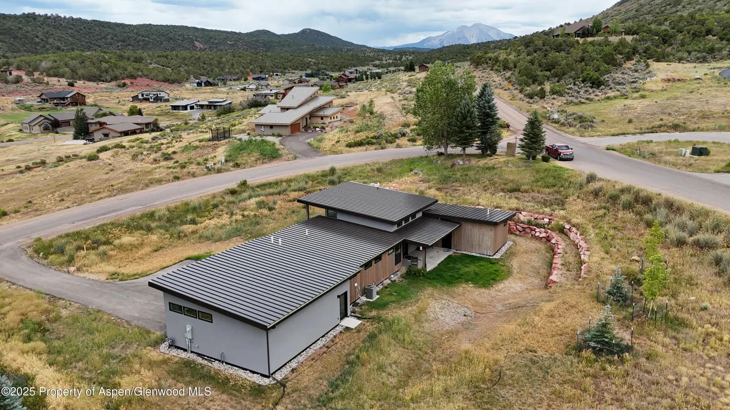 Aerial view of a modern gray house with a dark metal roof, surrounded by dry grass, trees, and mountains in the background.