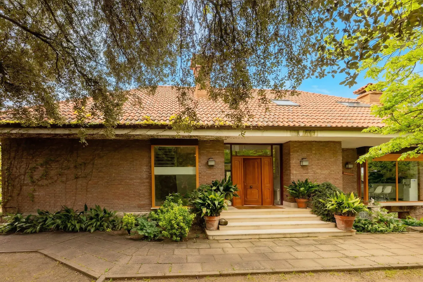 Exterior of a brick house with a red tile roof, a wooden door, and green plants.
