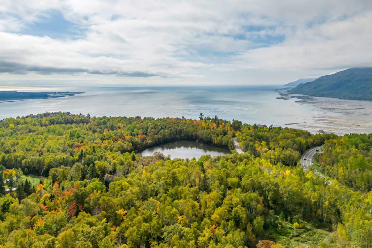 Aerial view of a lake surrounded by autumn trees, with a road winding along the coast and a bay in the background.