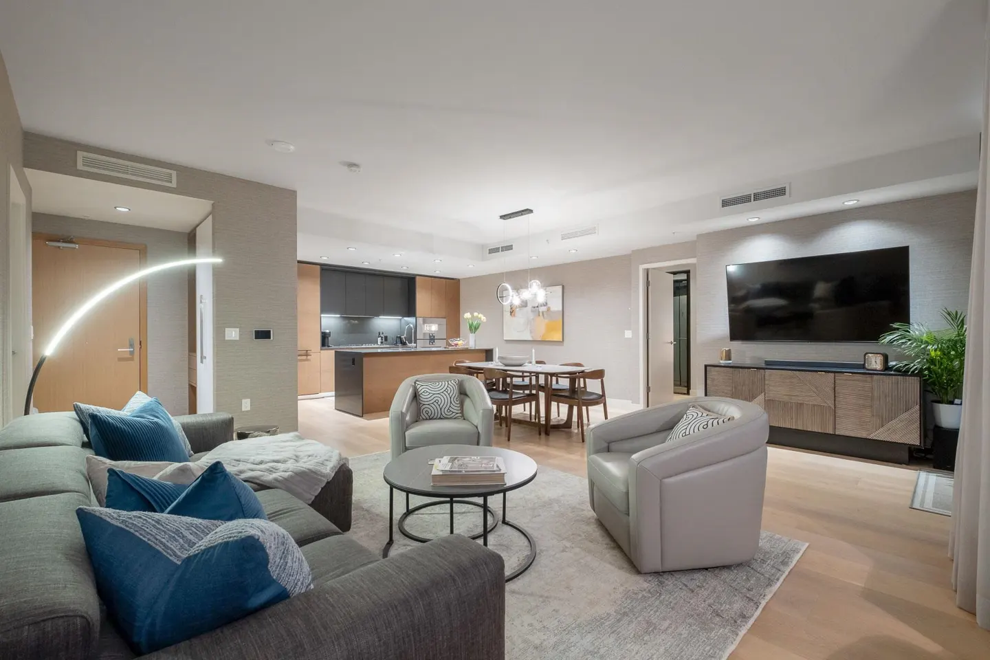 Open-concept living room with gray sofa, blue pillows, two gray chairs, and a round coffee table on a gray rug. Kitchen and dining area in background.