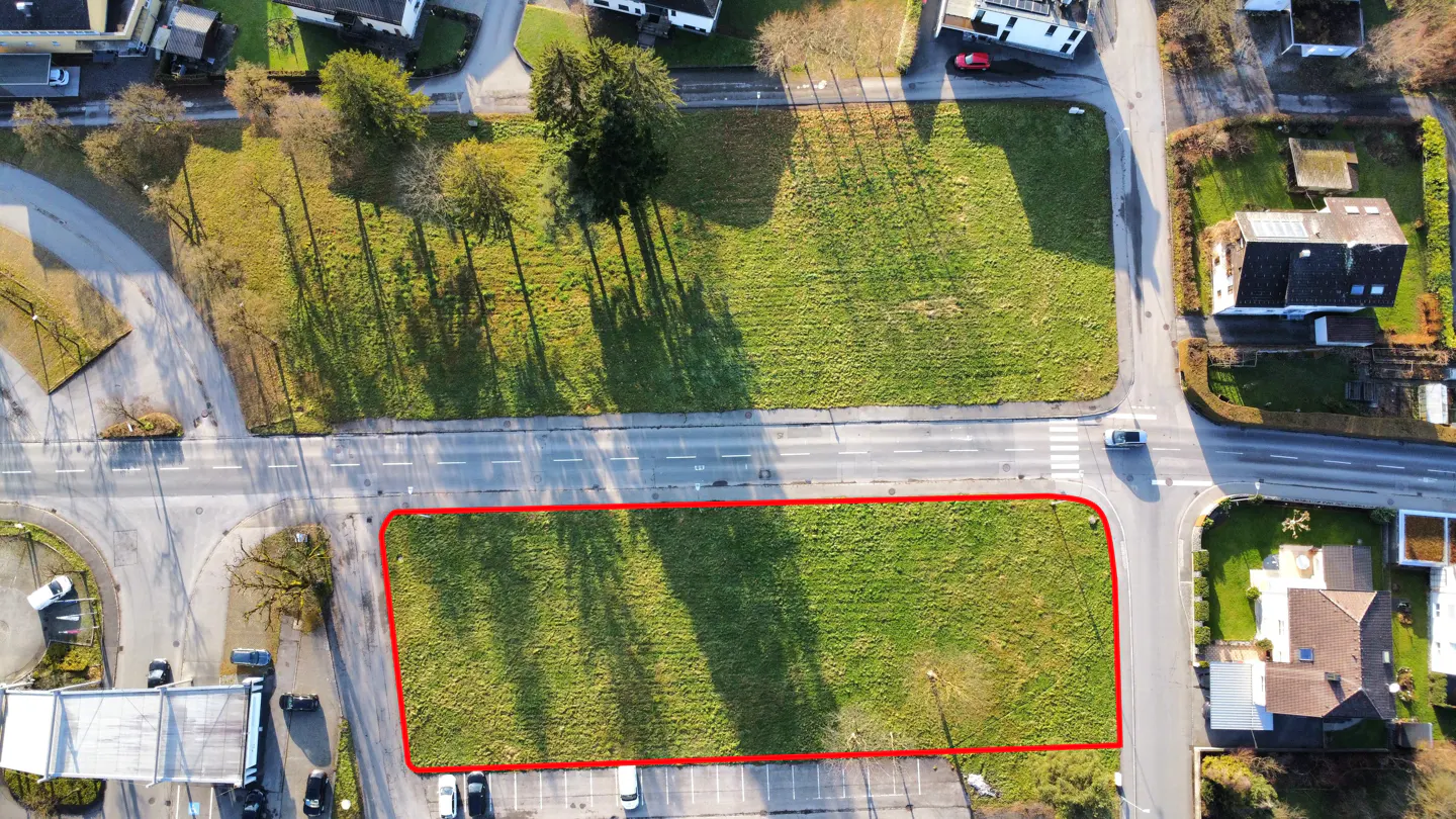 Aerial view of a grassy lot outlined in red, bordered by roads, trees, and buildings.