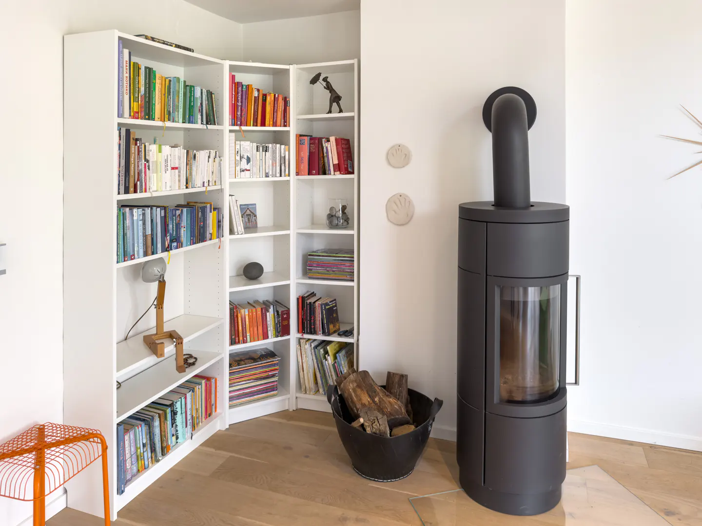 Bright room with white corner bookshelf filled with books, a black wood-burning stove, and a bucket of logs on a wood floor.