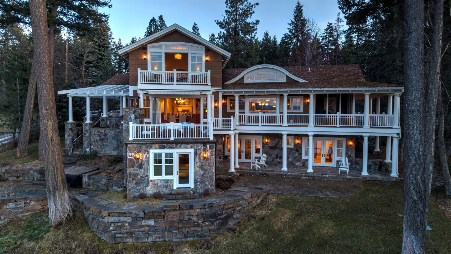 A multi-story brown house with white trim and stone accents, surrounded by trees. Balconies and lit windows add warmth.