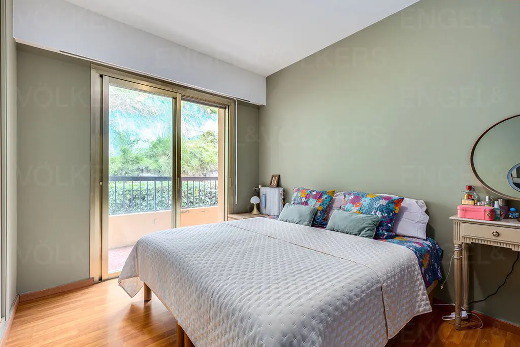 Bedroom with a white quilted bedspread, floral pillows, and a sliding glass door to a balcony with green foliage.