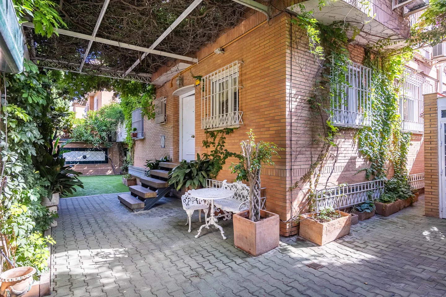 Brick house with a patio, vines, and a white metal table and chairs. The patio is paved with gray bricks. There are plants in pots around the patio.