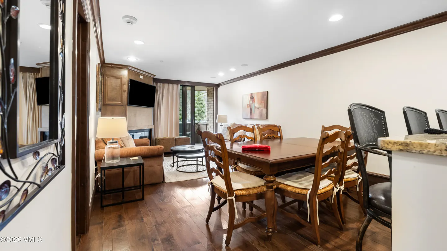 Interior view of a living and dining area with hardwood floors, a brown sofa, a wooden dining table with chairs, and a TV above a fireplace.