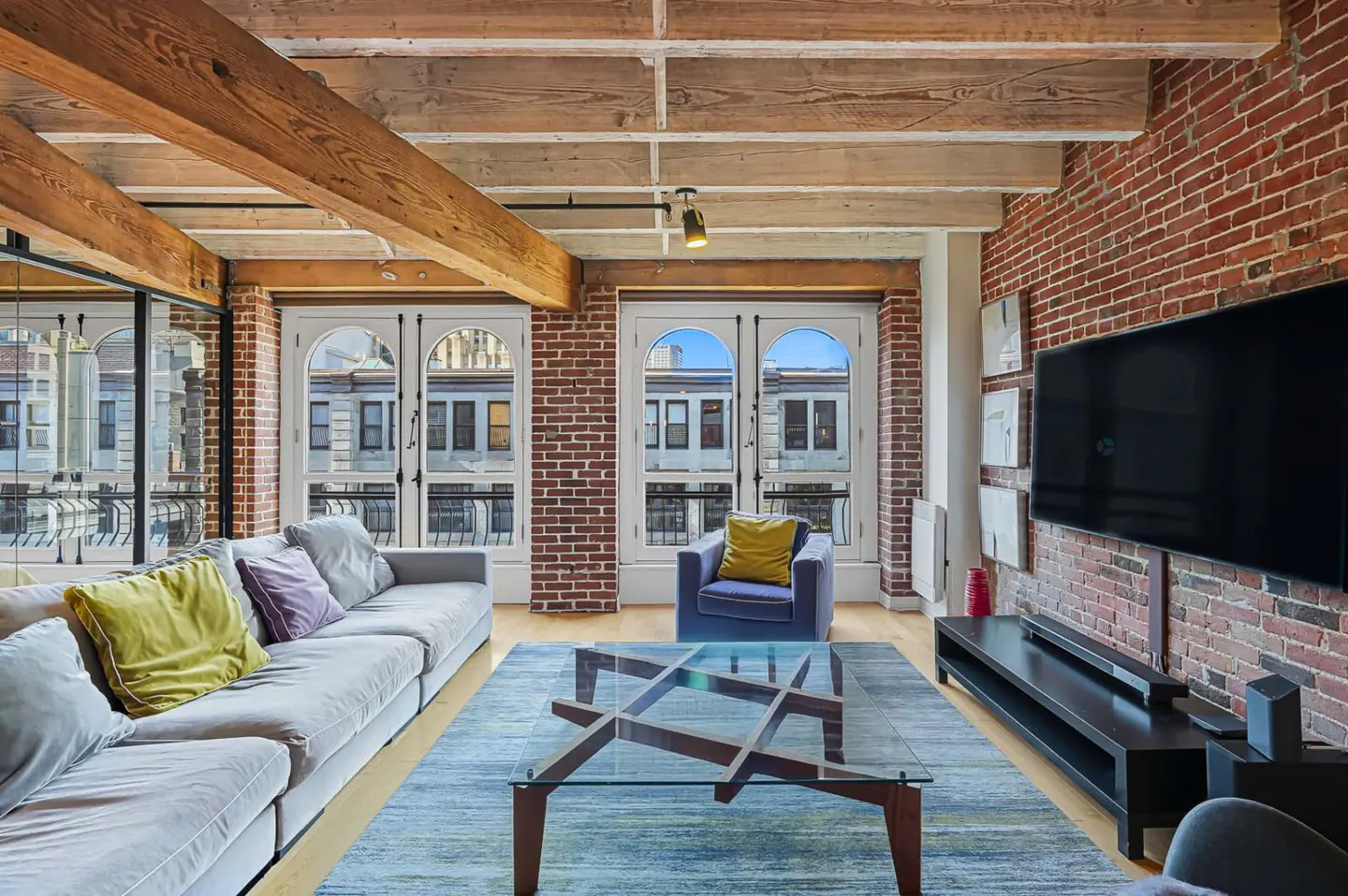 Loft apartment living room with exposed brick wall, wood beams, large windows, gray sofa, and blue rug.