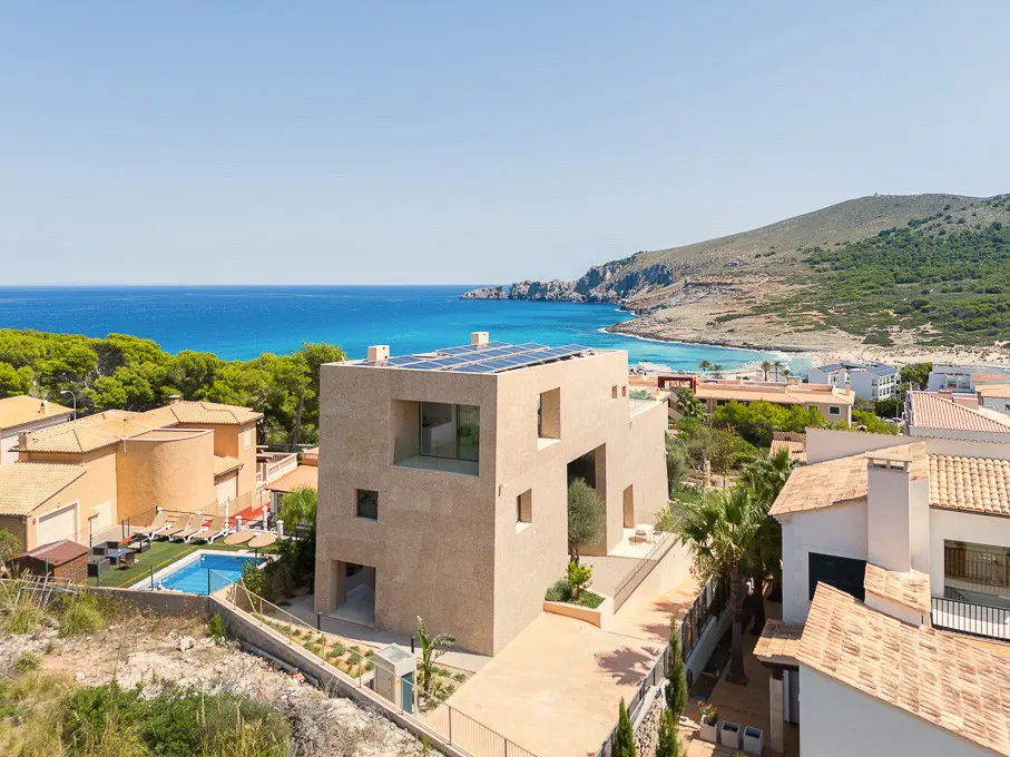 Modern beige villa with solar panels overlooking a blue Mediterranean bay, surrounded by coastal homes and pine trees.