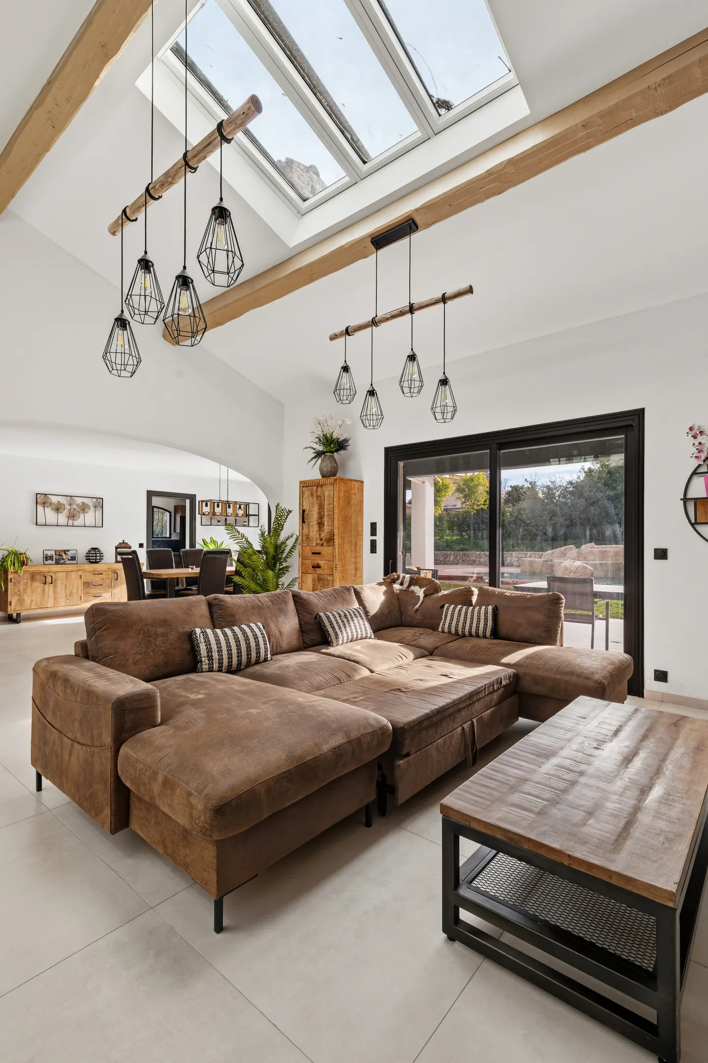 Bright living room with a large brown sectional sofa, wooden coffee table, and geometric pendant lights. Skylights and a sliding glass door provide natural light.