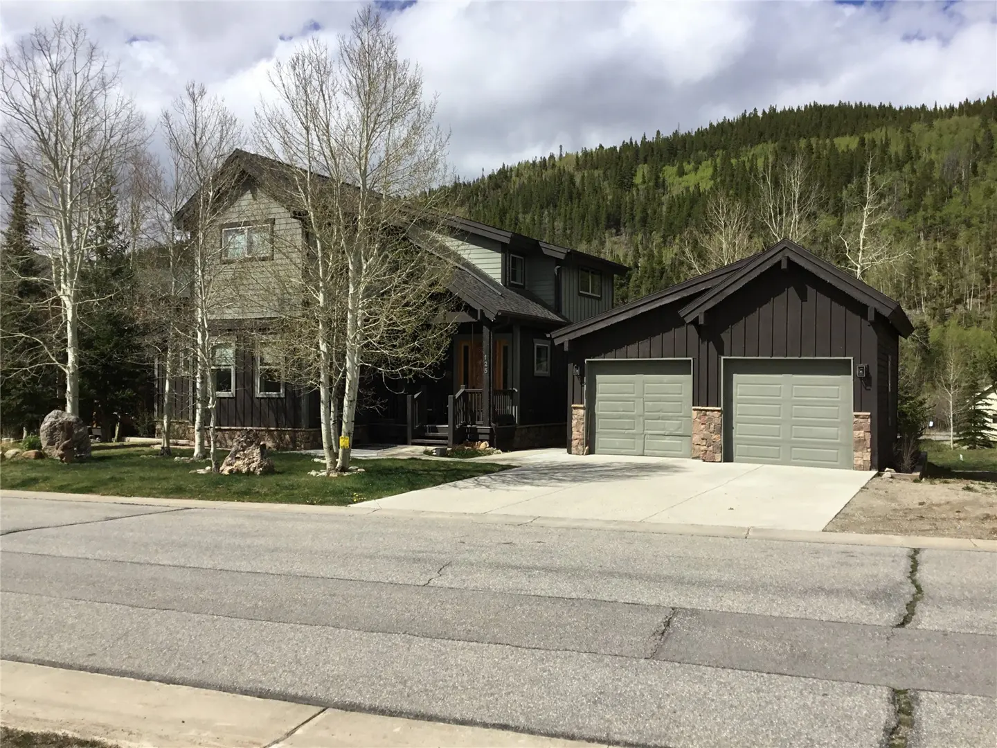 Two-story house with a two-car garage. The house is dark brown with light green accents. Trees and a mountain are in the background.