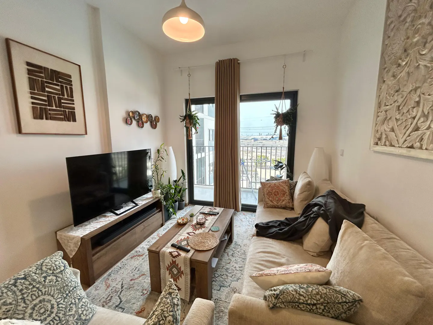 Living room with beige sofa, wooden coffee table, TV, and balcony view. Hanging plants and decorative wall art add a cozy touch.