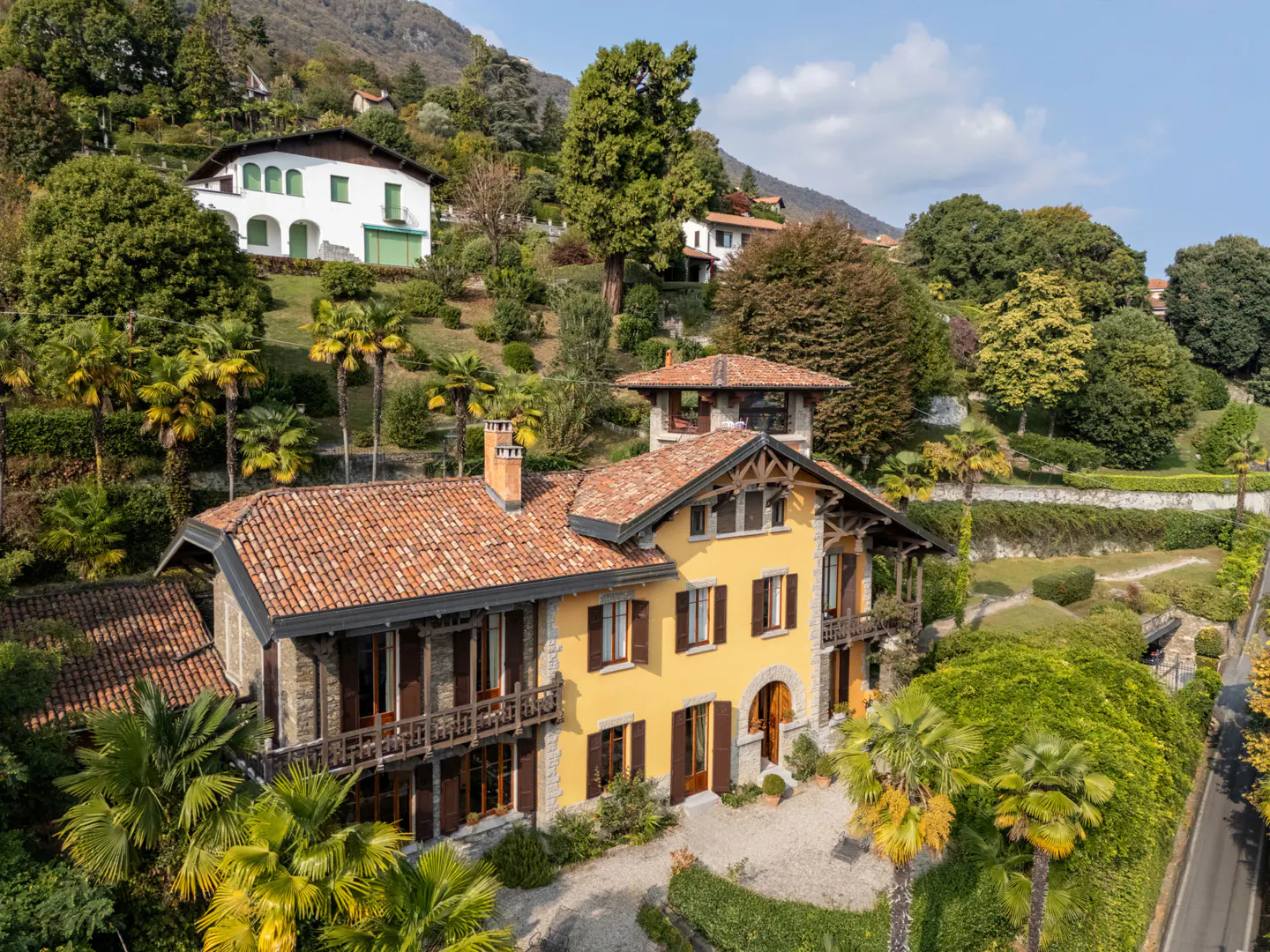 Aerial view of a yellow villa with a red tile roof, surrounded by lush greenery and palm trees on a hillside.