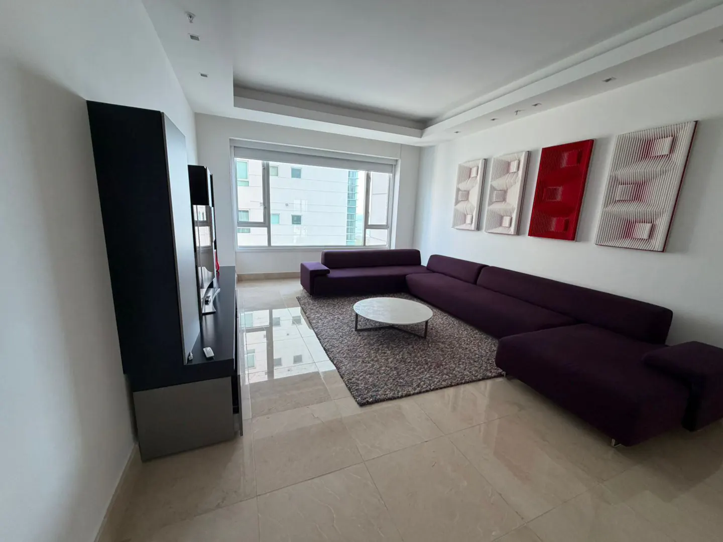 Bright living room with a purple sectional sofa, a white round table on a gray rug, and modern art on the wall.