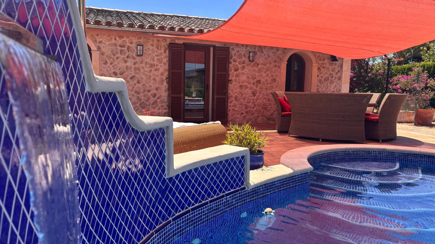 Outdoor patio with a pool, blue tile waterfall, and a stone building with a red awning. Wicker furniture is visible.