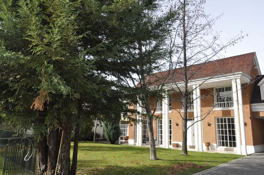 Brick house with white columns and a red roof, partially obscured by trees and a green lawn.