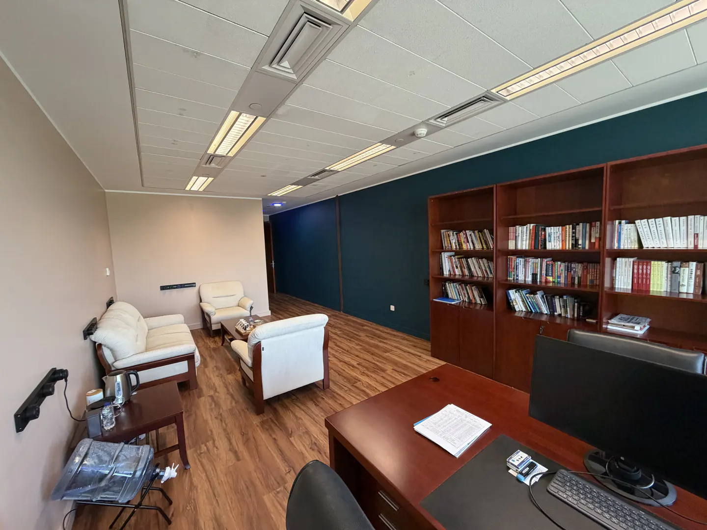 Office interior with white sofas, wooden floor, and a desk with a computer. A bookshelf filled with books stands against a dark blue wall.