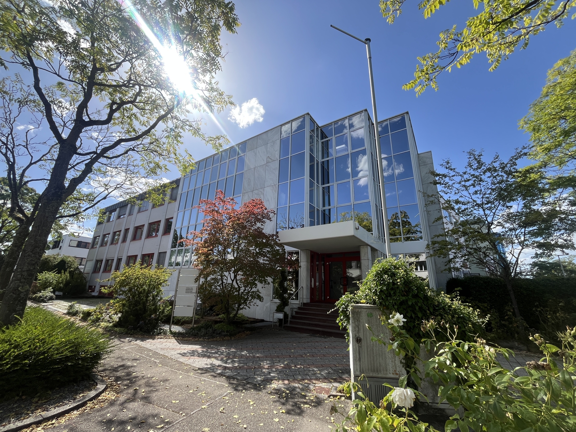 Modern office building with a glass facade, red entrance doors, and surrounding trees under a bright blue sky. [AI-generated text]