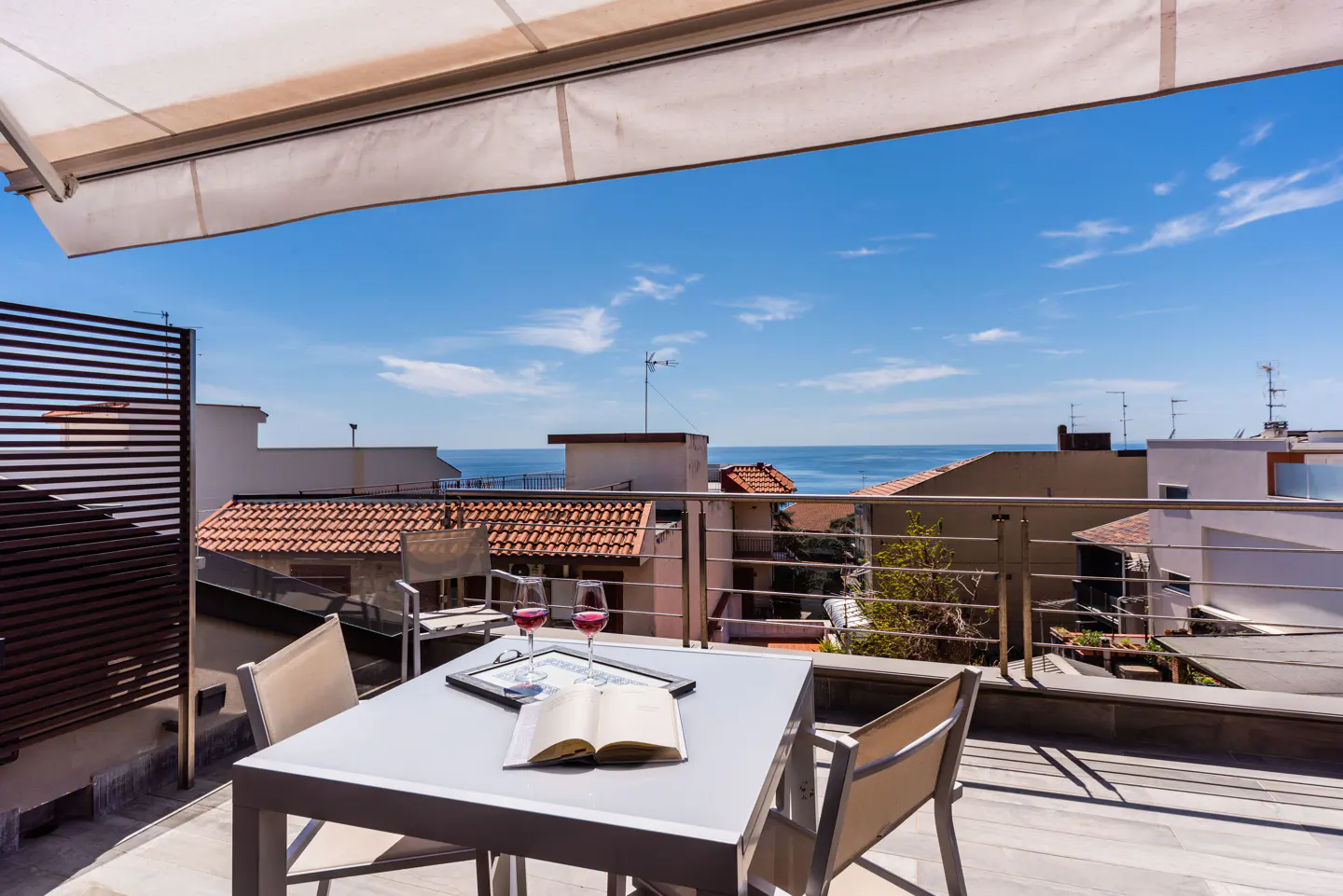 Balcony view with table, chairs, wine glasses, and open book. Ocean and rooftops in the background under a blue sky.