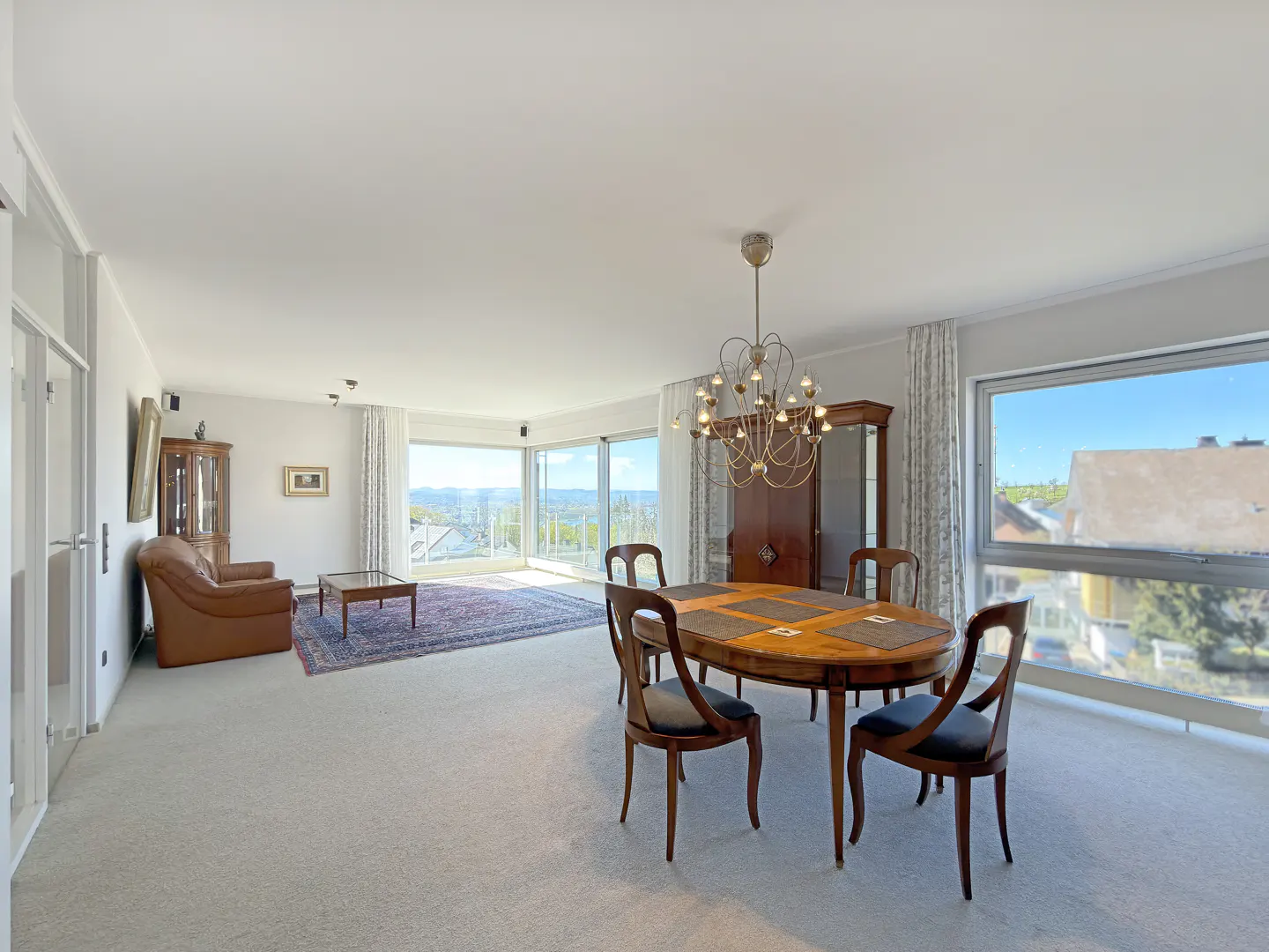Bright, open-plan living and dining area with large windows offering scenic views. Features a wooden dining table, chandelier, and comfortable seating area.