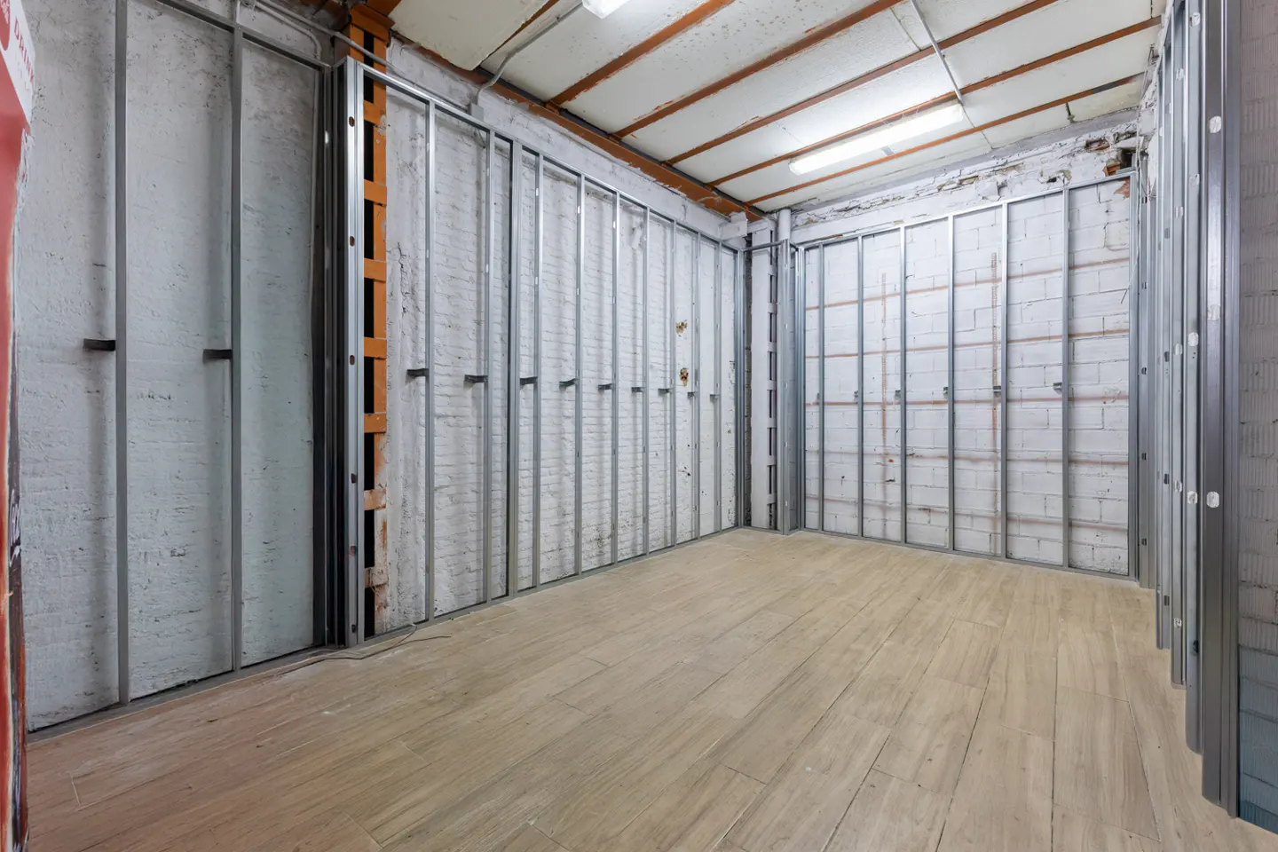 An unfinished room with metal wall studs over white brick walls and light wood-look tile flooring under fluorescent lights.
