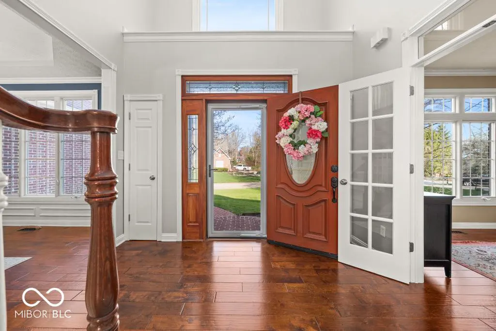 Open front door with floral wreath, wood floors, and white trim. View to the outside with green lawn and house.