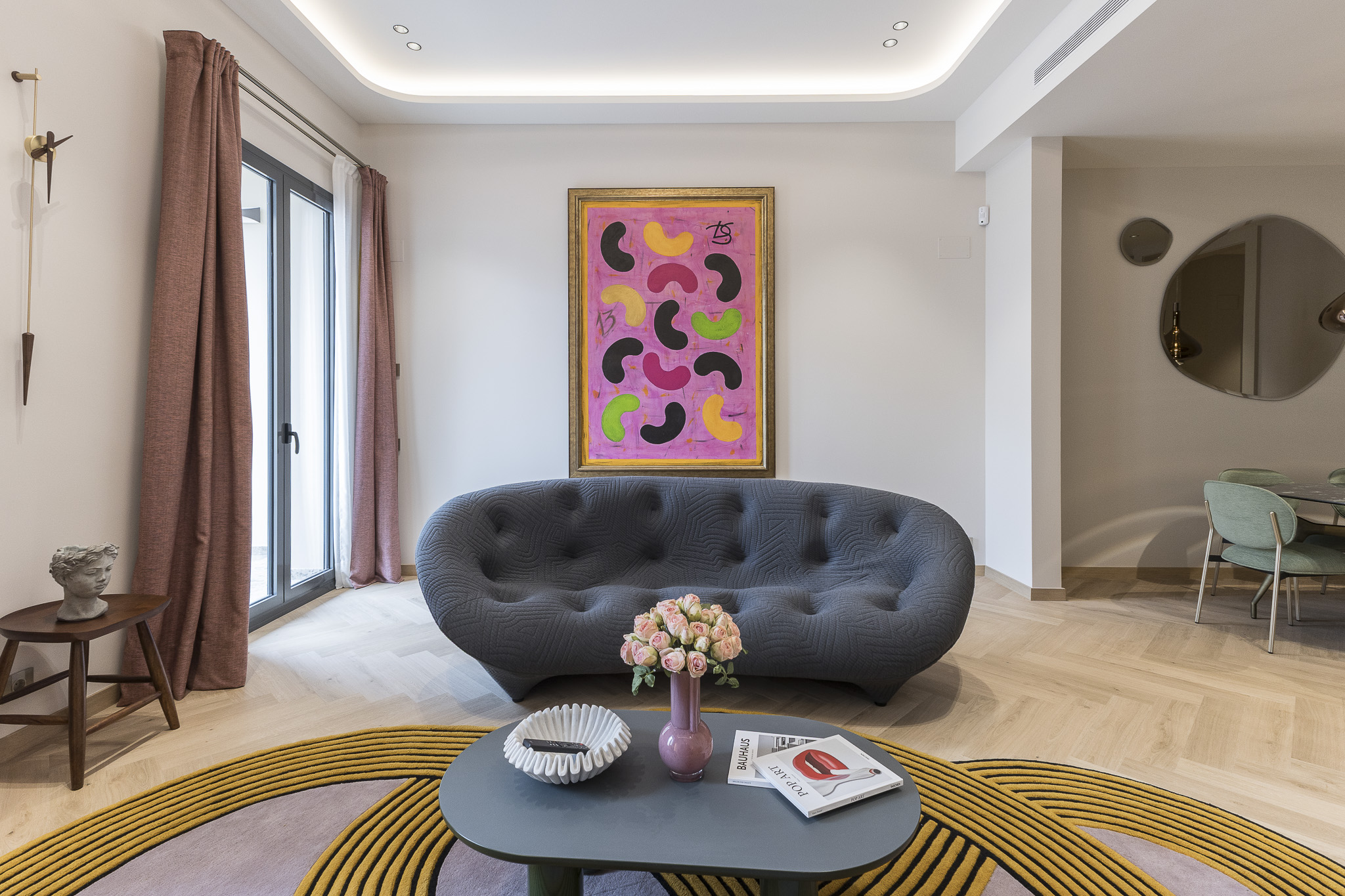 Living room with a gray sofa, pink abstract art, and a round rug with yellow lines. A coffee table holds flowers and magazines.