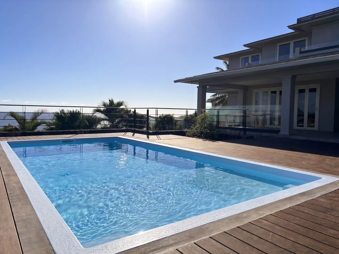 A rectangular pool with clear blue water sits on a wooden deck, with a house, ocean, and blue sky in the background.