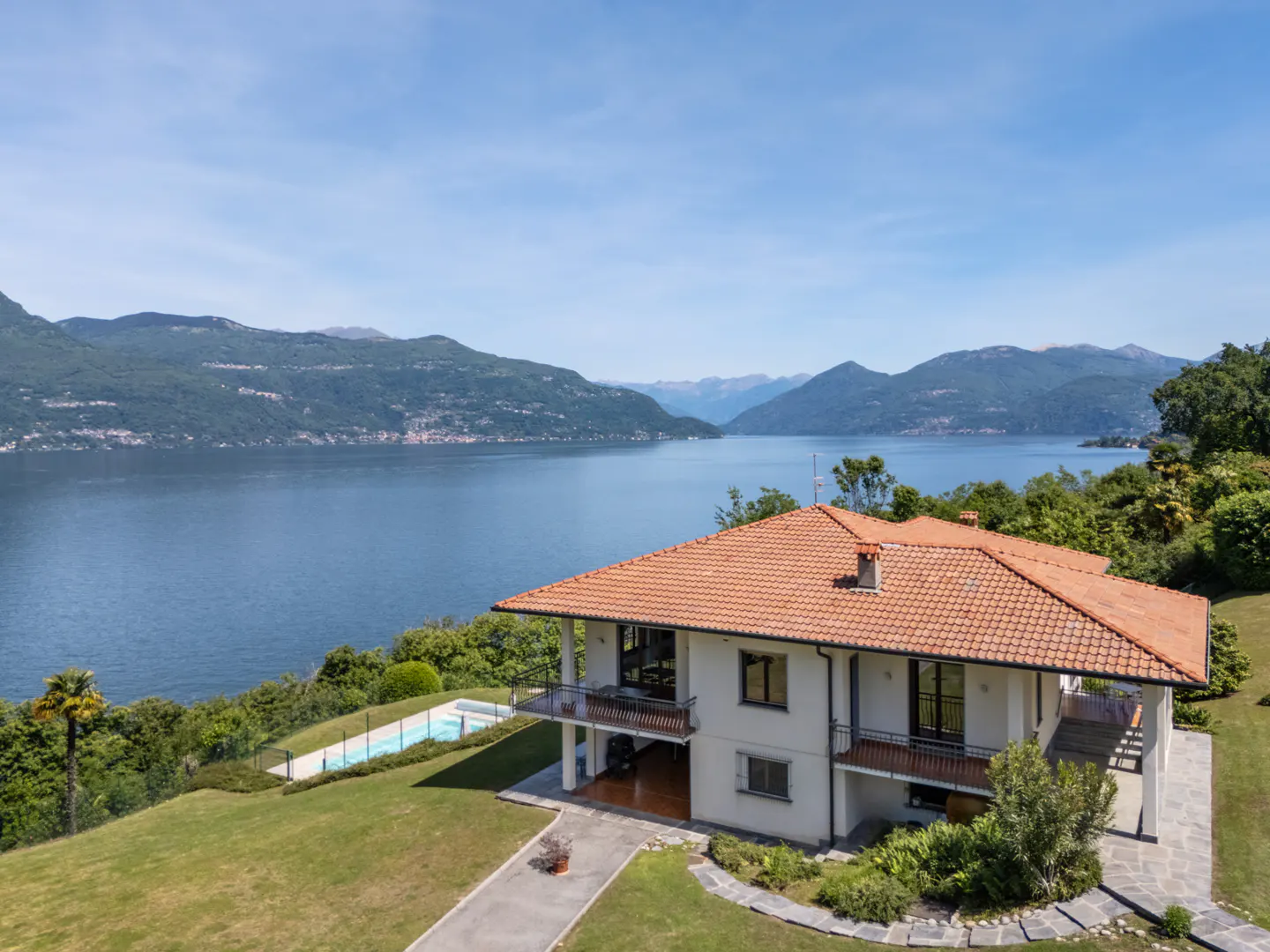 Two-story villa with a terracotta roof, pool, and lawn overlooking a large lake surrounded by mountains under a blue sky.