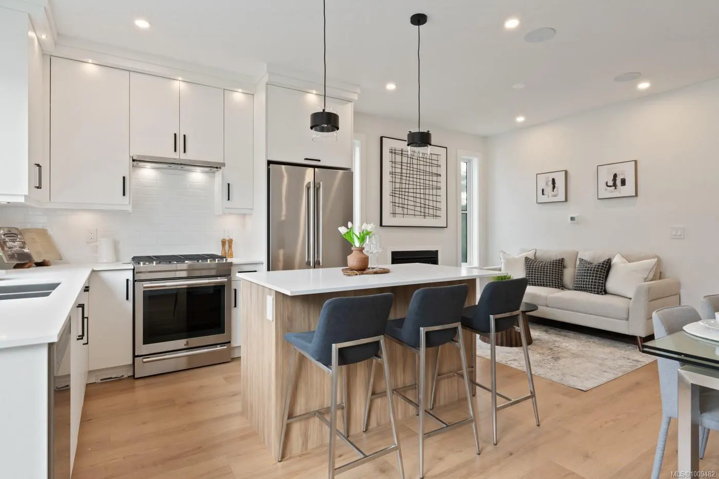 Bright, modern kitchen with white cabinets, stainless steel appliances, island with blue chairs, and living area with a beige sofa.