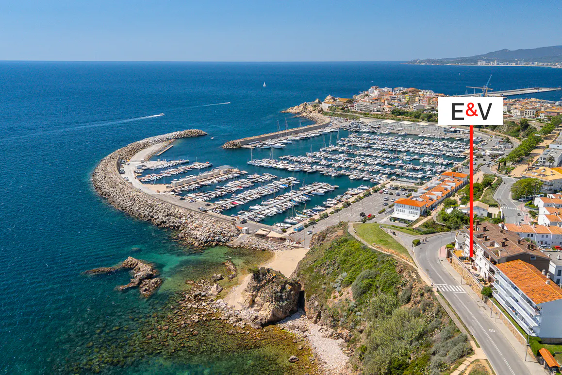 Aerial view of a marina filled with boats, next to a coastal town with a red line pointing to a building.