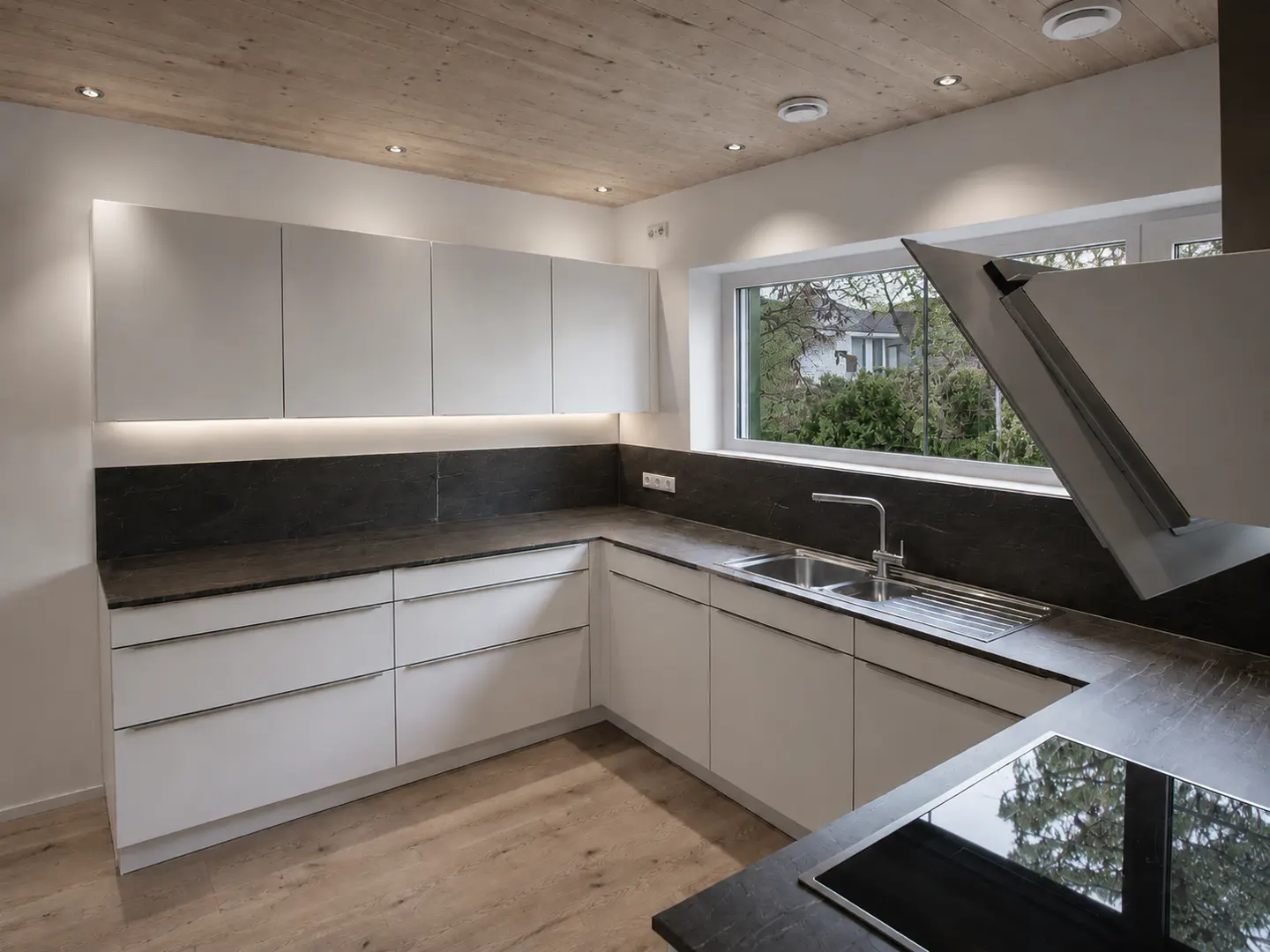 Bright kitchen with white cabinets, dark countertops, and a wood ceiling. A window overlooks a green yard.