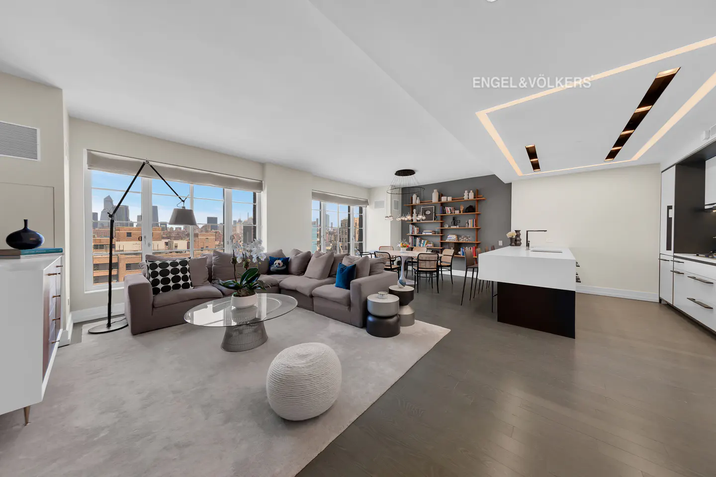 Open-concept living space with a gray sectional sofa, glass coffee table, and city views from large windows. A dining area and kitchen are visible.