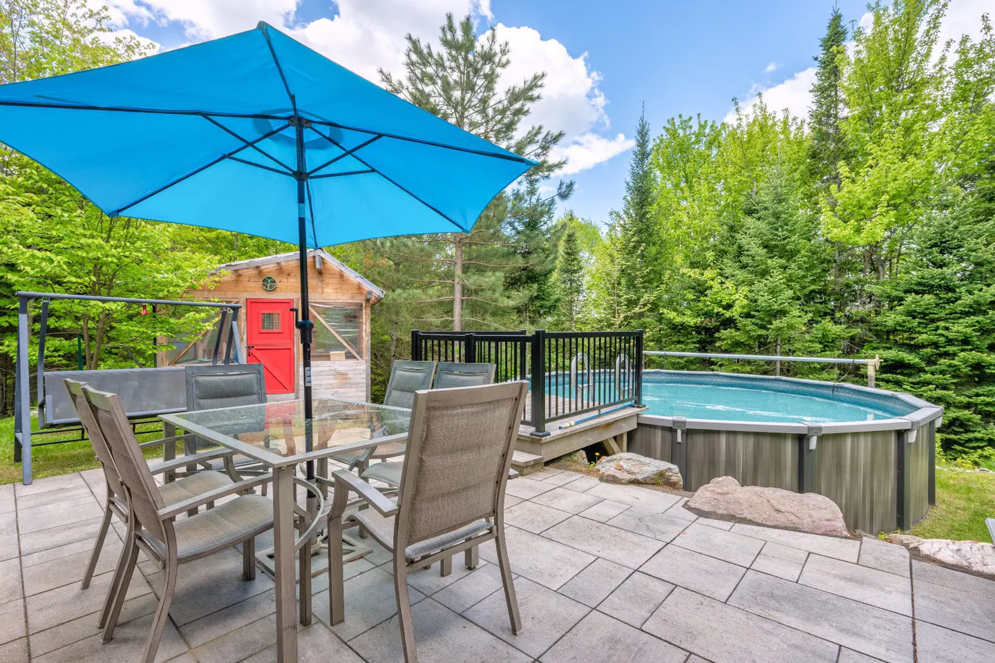 Patio with a glass table, blue umbrella, and chairs next to an above-ground pool in a lush, wooded backyard.