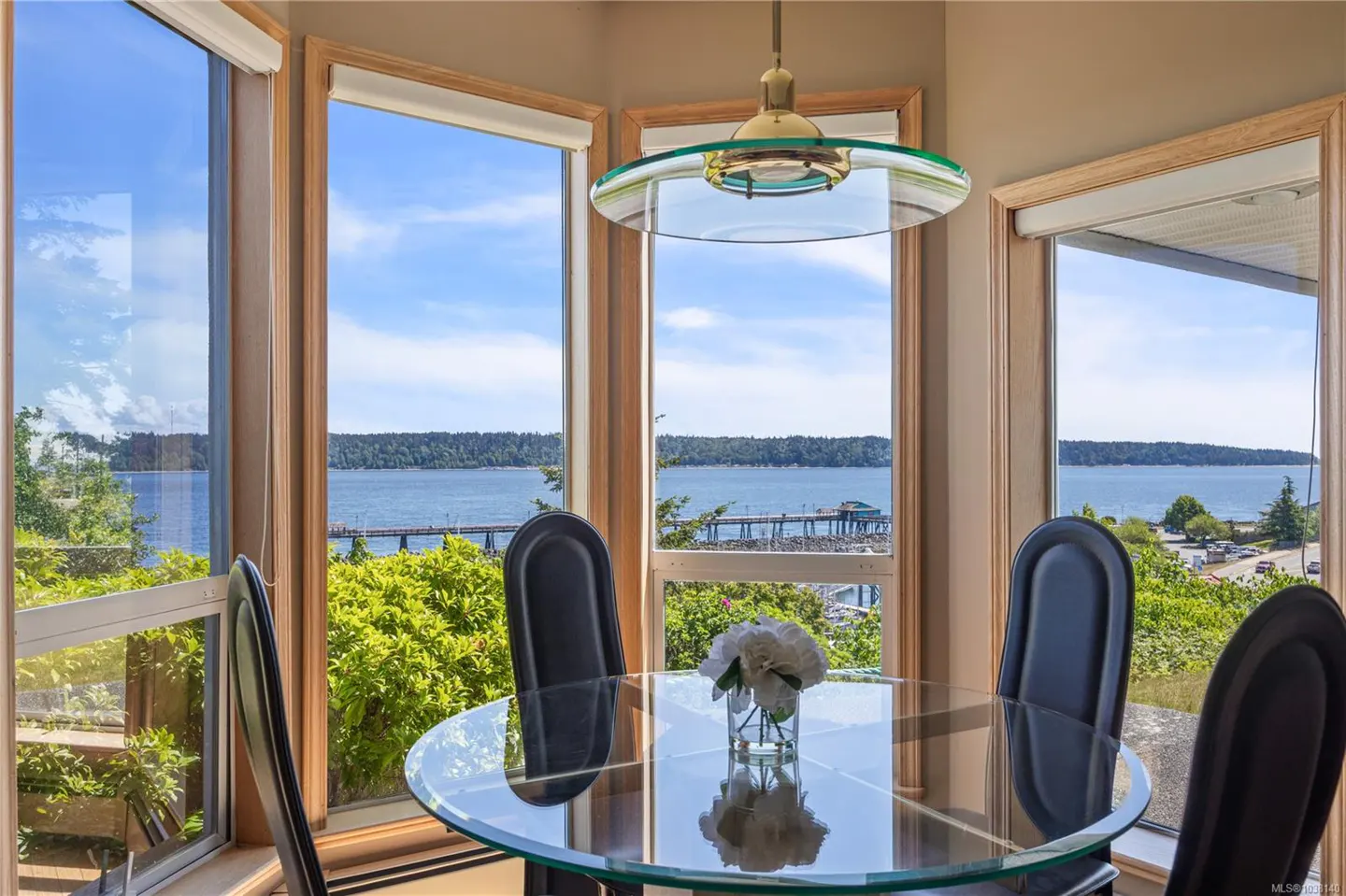 A dining area with a glass table, black chairs, and a view of the ocean through large windows. A glass pendant light hangs above the table.