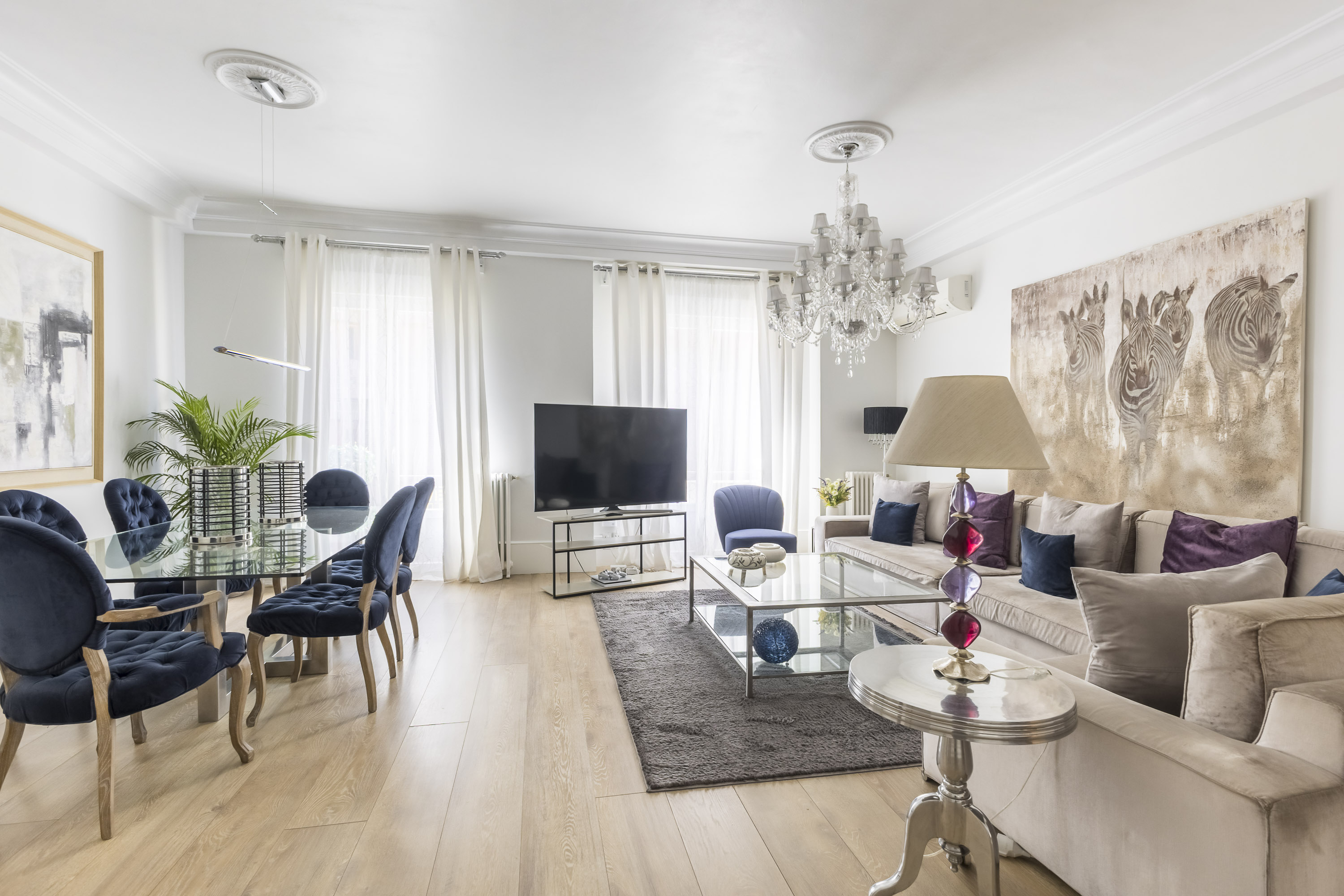 Elegant living and dining room with a crystal chandelier, navy velvet chairs, beige sofa, and zebra wall art. [AI-generated text]