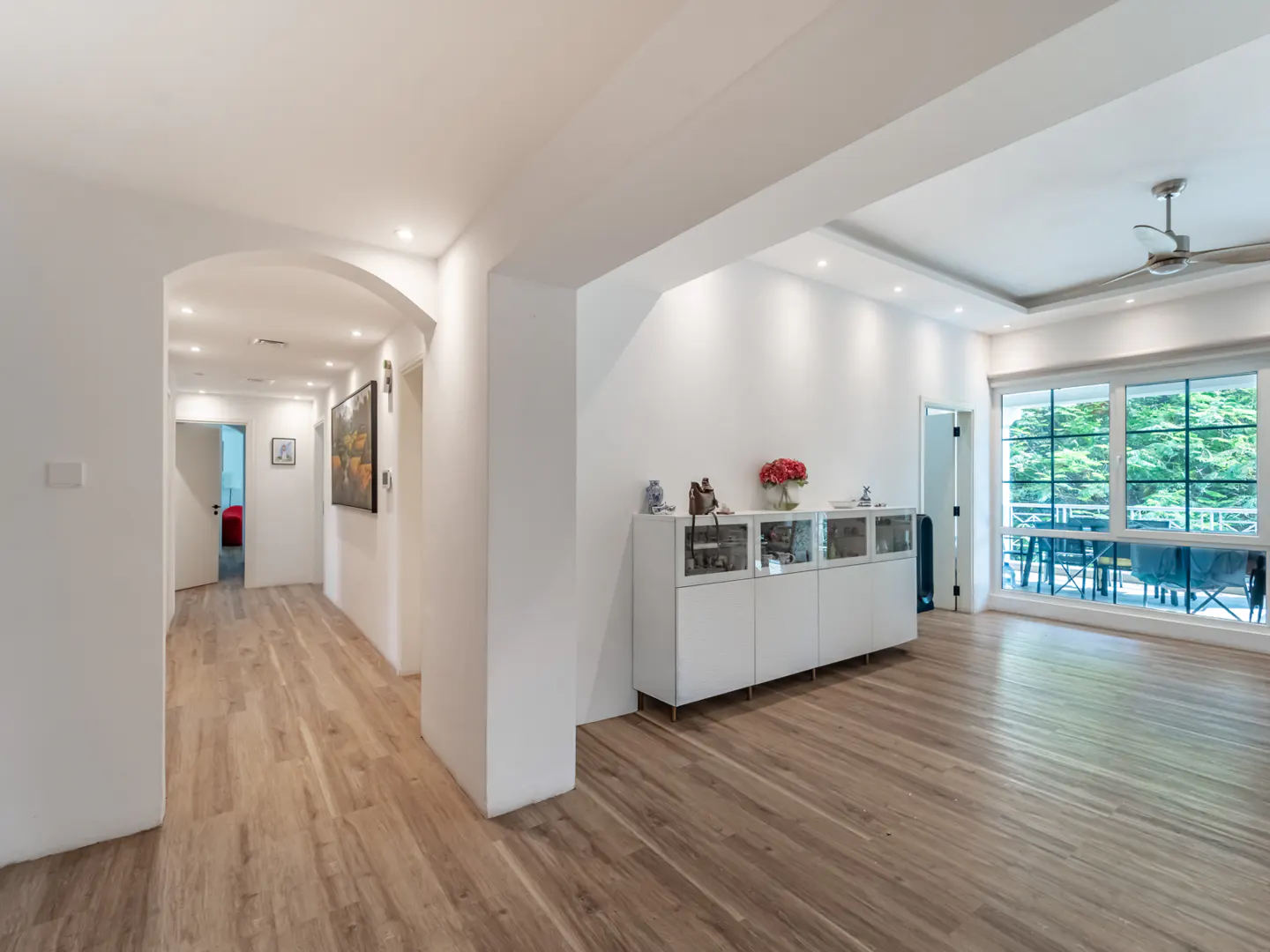 Bright, open-concept home interior with wood floors, white walls, and recessed lighting. A hallway leads to a room with red accents. A cabinet sits near a window with a patio view.
