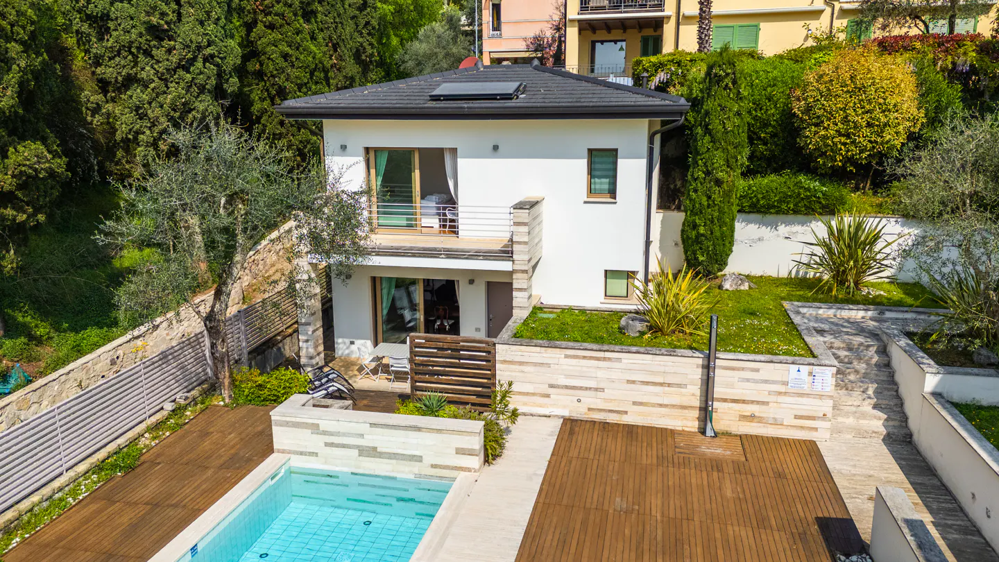 Two-story white house with a pool and wooden deck surrounded by green trees and plants.