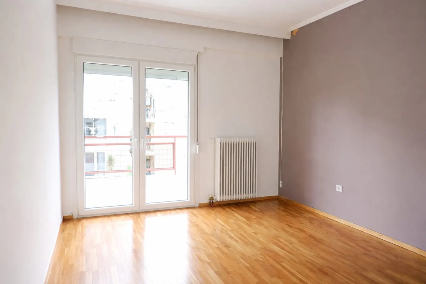 Bright, empty room with hardwood floors, white walls, and a gray accent wall. A glass door leads to a balcony. A white radiator is on the wall.