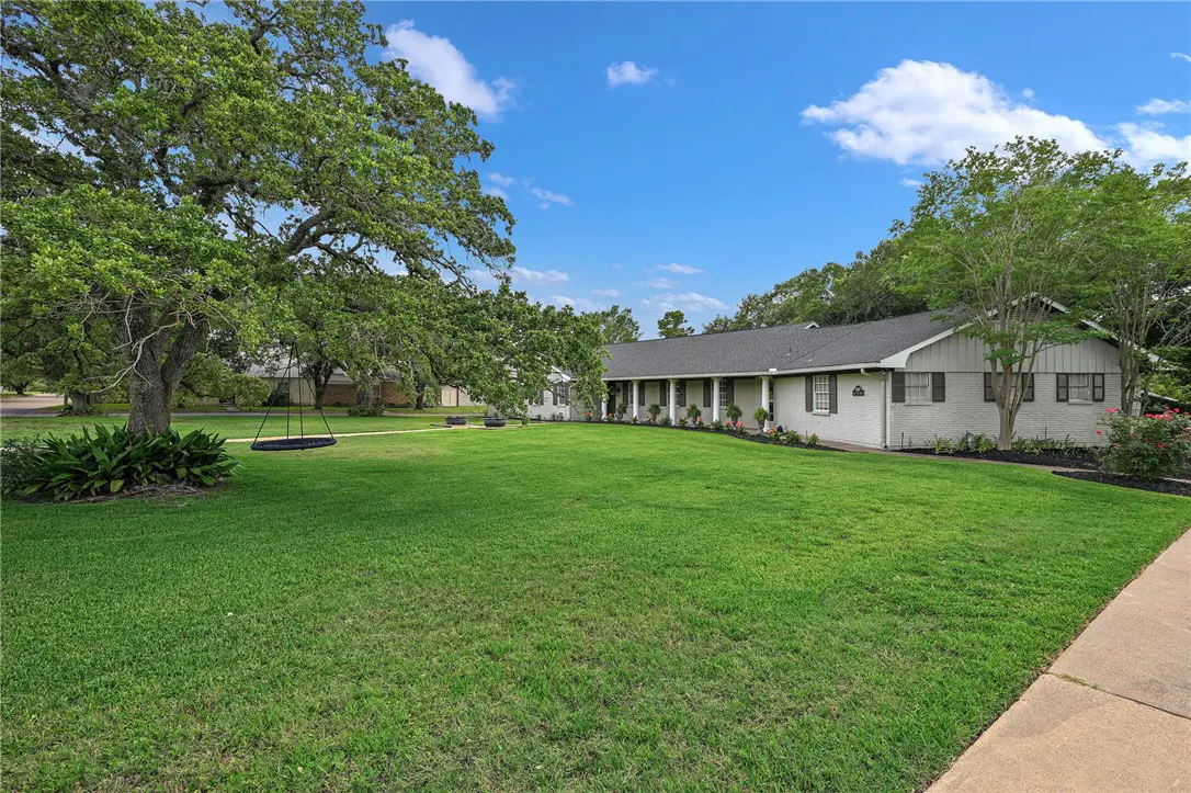 Single-story white ranch house with a large green lawn, mature trees, and a hanging swing under a blue sky.