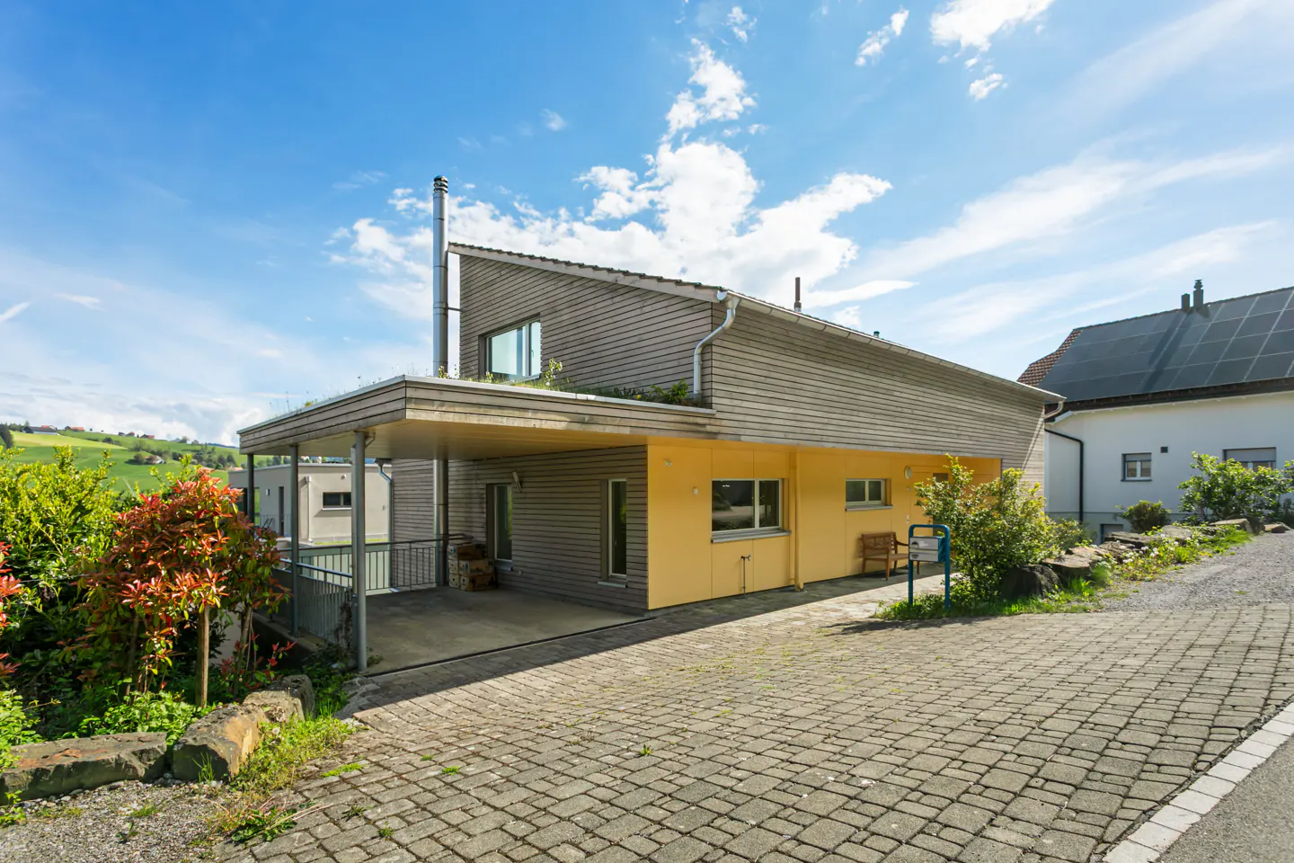 Exterior view of a modern two-story house with wood siding and a yellow accent wall, featuring a carport and a paved driveway.