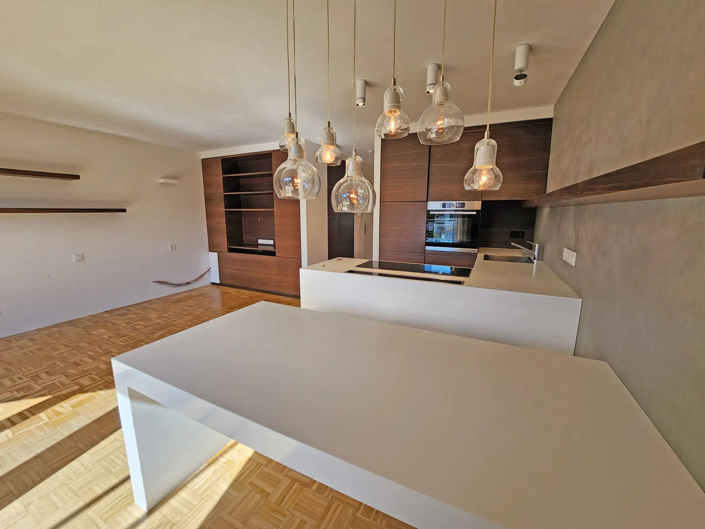 A modern kitchen with a white island, wood floors, and brown cabinets. Hanging glass pendant lights illuminate the space.
