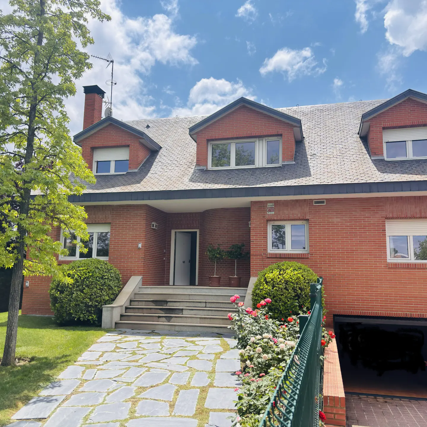 Red brick house with dormer windows, gray roof, and stone walkway leading to the front door. Green lawn and trees surround the house.