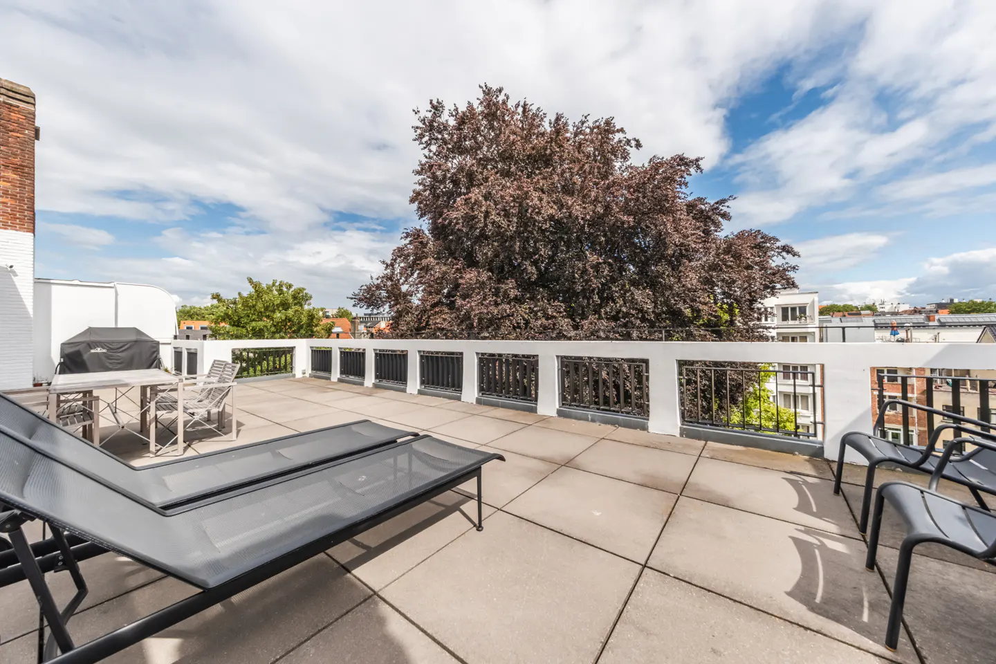 Spacious rooftop terrace with tiled flooring, lounge chairs, dining set, and a white railing overlooking city trees.