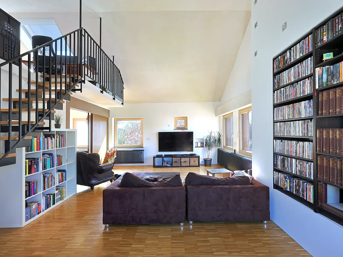 Bright living room with a brown sofa, TV, and a black staircase leading to a loft with a black railing. A large bookshelf is on the right.