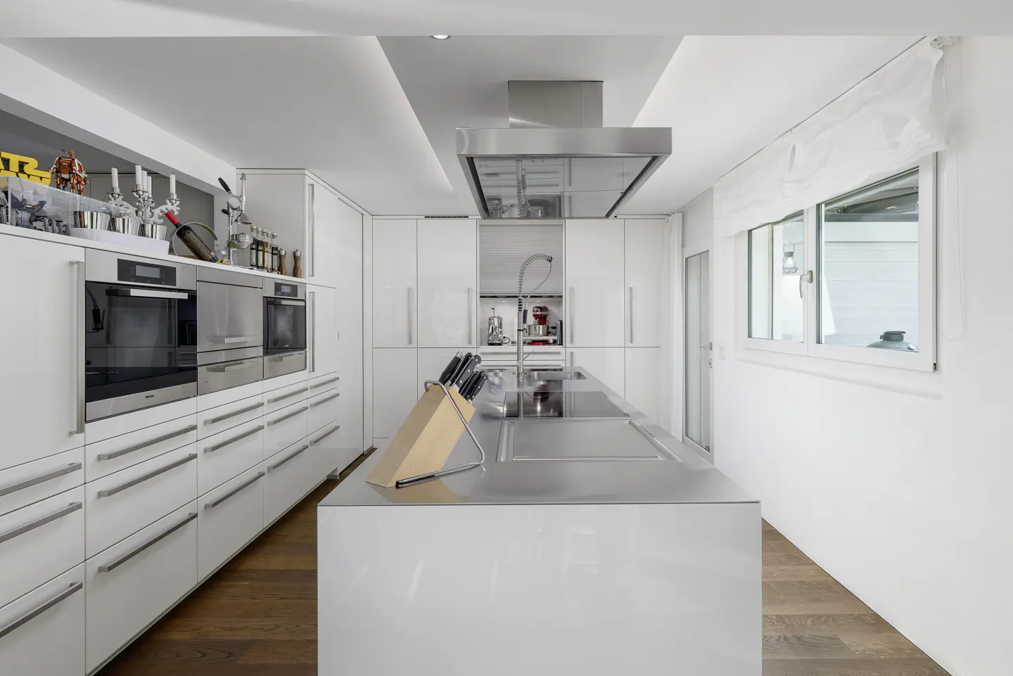 A modern kitchen with white cabinets, stainless steel island, and wood floors. A window with a white curtain is on the right.