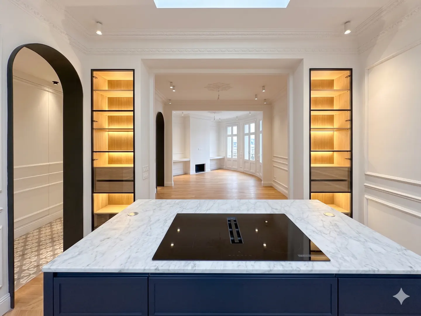 Modern kitchen island with marble countertop and black cooktop, facing a bright living area with arched doorways and shelves.
